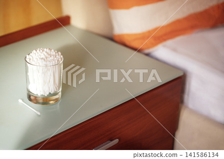 Glass container filled with cotton swabs placed on a modern wooden nightstand beside a bed with orange striped pillow and soft bedding visible in the background Glass container filled with cotton swabs placed on a modern wooden nightstand beside a bed with orange striped pillow and soft bedding visible in the background 141586134