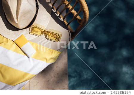 Summer poolside scene featuring a straw hat, yellow sunglasses, and sunscreen on a striped towel beside a clear blue swimming pool Summer poolside scene featuring a straw hat, yellow sunglasses, and sunscreen on a striped towel beside a clear blue swimming pool 141586154
