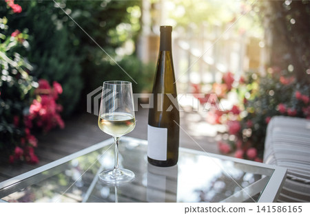 Glass of white wine beside a bottle on a glass table in a garden setting with colorful flowers and greenery in the background during a sunny day Glass of white wine beside a bottle on a glass table in a garden setting with colorful flowers and greenery in the background during a sunny day 141586165