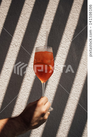 Hand holding a tall glass of orange cocktail with ice and straw, illuminated by sunlight casting striped shadows on a textured wall background Hand holding a tall glass of orange cocktail with ice and straw, illuminated by sunlight casting striped shadows on a textured wall background 141586166