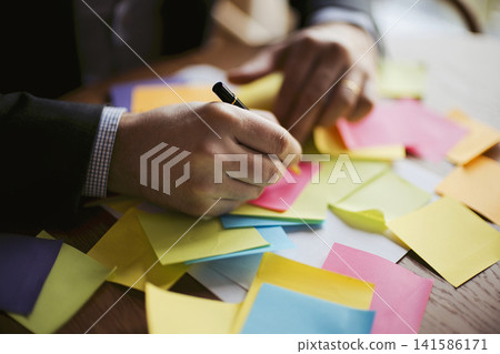 Male office worker in a suit writing on a sticky note surrounded by colorful notes on a wooden desk, emphasizing organization and brainstorming activities Male office worker in a suit writing on a sticky note surrounded by colorful notes on a wooden desk, emphasizing organization and brainstorming activities 141586171