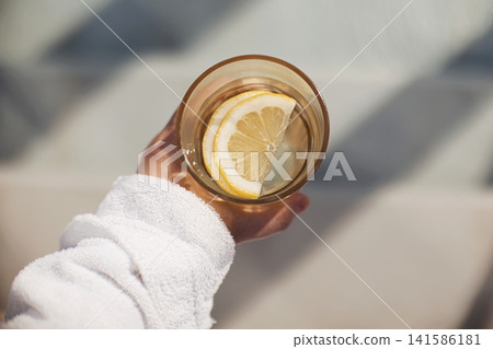 Hand holding a glass of water with a lemon slice, wearing a white towel, sunlight casting shadows on a light surface in a serene setting Hand holding a glass of water with a lemon slice, wearing a white towel, sunlight casting shadows on a light surface in a serene setting 141586181