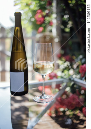 White wine bottle and glass filled with white wine placed on a glass table surrounded by blooming flowers in a sunny outdoor setting White wine bottle and glass filled with white wine placed on a glass table surrounded by blooming flowers in a sunny outdoor setting 141586183