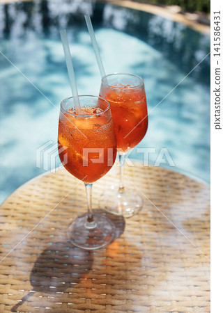 Two elegant glasses of orange cocktail with ice and straws placed on a wicker table beside a sparkling blue swimming pool under bright sunlight Two elegant glasses of orange cocktail with ice and straws placed on a wicker table beside a sparkling blue swimming pool under bright sunlight 141586431