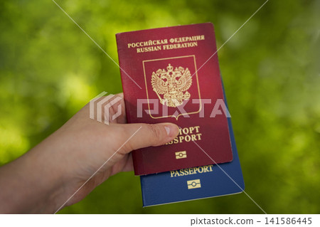 Hand holding two passports, one red Russian Federation passport and one blue passport, with blurred green background indicating outdoor setting 141586445