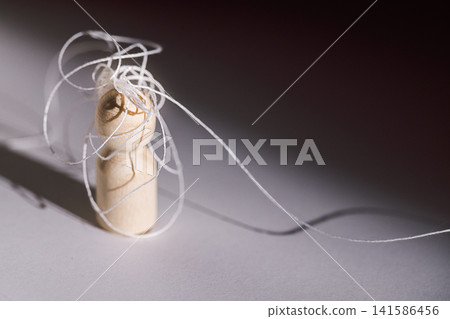 Wooden figurine with tangled white thread on a gray surface, showcasing intricate details and shadows in a minimalist composition Wooden figurine with tangled white thread on a gray surface, showcasing intricate details and shadows in a minimalist composition 141586456