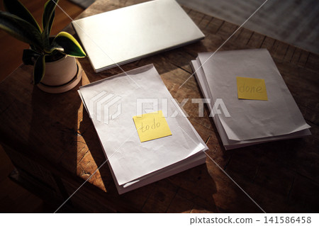 Workspace with organized papers labeled 'todo' and 'done', a laptop and a potted plant on a wooden table in a well-lit indoor environment Workspace with organized papers labeled 'todo' and 'done', a laptop and a potted plant on a wooden table in a well-lit indoor environment 141586458
