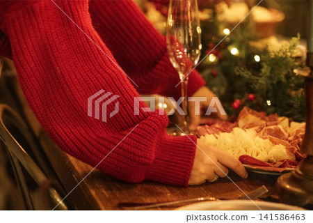 Hands of a person in a red sweater arranging a festive charcuterie board with assorted meats and cheeses on a wooden table with holiday decorations and glassware Hands of a person in a red sweater arranging a festive charcuterie board with assorted meats and cheeses on a wooden table with holiday decorations and glassware 141586463