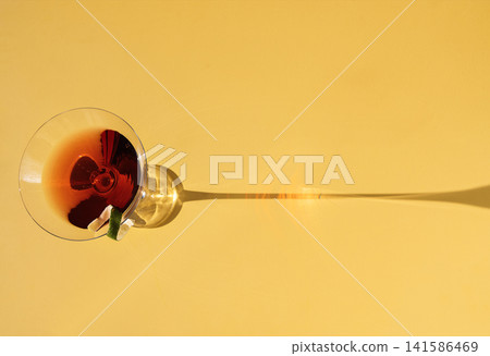 Close-up of a martini glass on a yellow background. View from above. An elegant goblet with a red alcoholic sparkling drink decorated with lime zest. Sunlight. Close-up of a martini glass on a yellow background. View from above. An elegant goblet with a red alcoholic sparkling drink decorated with lime zest. Sunlight. 141586469