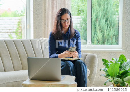 Focused businesswoman sitting on sofa using smartphone while working on laptop at modern cozy home Focused businesswoman sitting on sofa using smartphone while working on laptop at modern cozy home 141586915