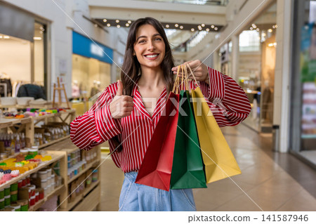 Smiling young woman feeling happy and contented while doing shopping in a shopping center Smiling young woman feeling happy and contented while doing shopping in a shopping center 141587946