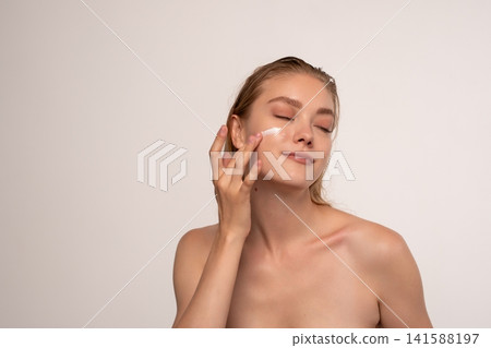 Young woman applying facial cream on cheek, enjoying her skincare routine for healthy, glowing skin Young woman applying facial cream on cheek, enjoying her skincare routine for healthy, glowing skin 141588197