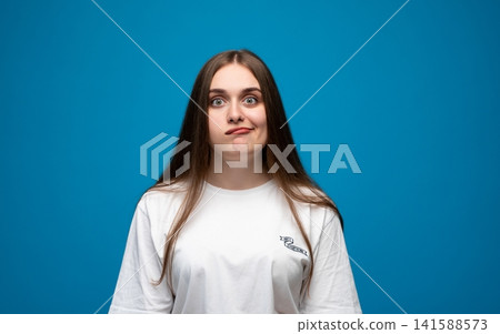 Young woman expressing confusion and uncertainty, having doubt, looking perplexed in studio portrait against blue background Young woman expressing confusion and uncertainty, having doubt, looking perplexed in studio portrait against blue background 141588573