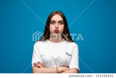 Young woman pouting with pursed lips and crossed arms, showing annoyance or playful frustration on blue background Young woman pouting with pursed lips and crossed arms, showing annoyance or playful frustration on blue background 141588581