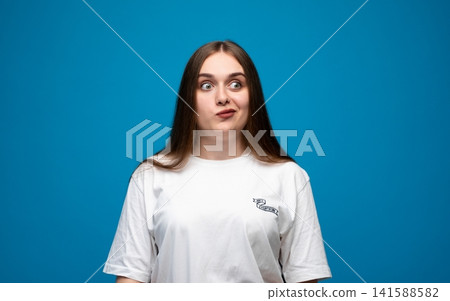 Young woman having an unsure and curious facial expression, looking away with doubt, standing against a blue background Young woman having an unsure and curious facial expression, looking away with doubt, standing against a blue background 141588582