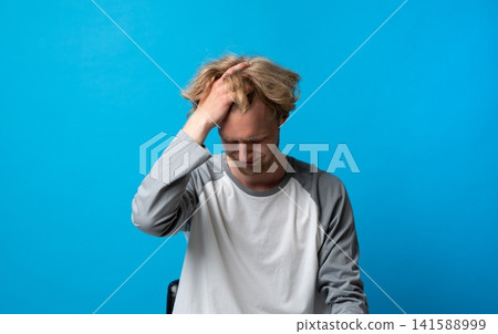 Young man experiencing severe headache, stress, and frustration, grimacing and holding his head in discomfort, suffering from pain and mental distress on blue background 141588999