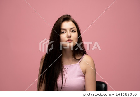 Young woman with serious expression posing against a vibrant pink background, long dark hair falling over shoulders Young woman with serious expression posing against a vibrant pink background, long dark hair falling over shoulders 141589010