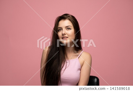 Young woman with long dark hair wearing a pink sleeveless top, posing on a pink background, looking away Young woman with long dark hair wearing a pink sleeveless top, posing on a pink background, looking away 141589018