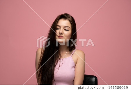 Young brunette woman sitting with long dark hair, looking down with a subtle smile on a pink background 141589019