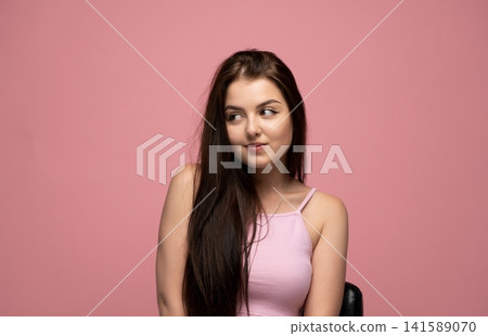 Young brunette woman looking sideways with shy smile on pink background, feeling modesty and introversion Young brunette woman looking sideways with shy smile on pink background, feeling modesty and introversion 141589070
