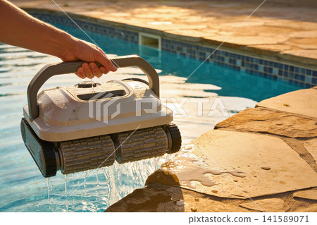 Hand lifting a robotic pool cleaner out of the water near the pool edge 141589071