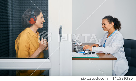 Audiological exam. Audiologist doing hearing check-up to a senior citizen woman in soundproof audiometric booth. Hearing loss treatment, side view Audiological exam. Audiologist doing hearing check-up to a senior citizen woman in soundproof audiometric booth. Hearing loss treatment, side view 141589380