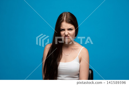 Young woman showing strong disgust, grimacing with a displeased and upset facial expression on blue background Young woman showing strong disgust, grimacing with a displeased and upset facial expression on blue background 141589396