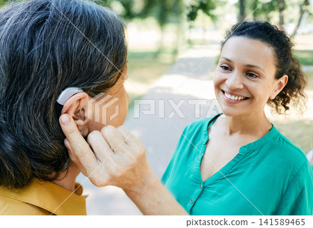 Mature woman with a hearing impairment uses a hearing aid to communicate with her female friend outdoor. Hearing solutions Mature woman with a hearing impairment uses a hearing aid to communicate with her female friend outdoor. Hearing solutions 141589465