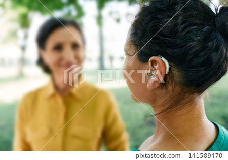 Adult woman with a hearing impairment uses a hearing aid to communicate with her female friend at city park. Hearing solutions Adult woman with a hearing impairment uses a hearing aid to communicate with her female friend at city park. Hearing solutions 141589470