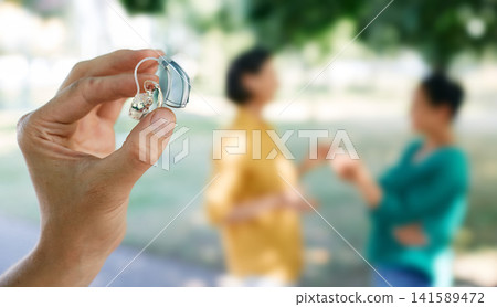 Hearing solutions. Last generation of hearing aid device in human hand with unfocused talking people in background. Full life with hearing aids Hearing solutions. Last generation of hearing aid device in human hand with unfocused talking people in background. Full life with hearing aids 141589472