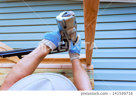Handy worker is fastening wooden boards with air nail gun on building site Handy worker is fastening wooden boards with air nail gun on building site 141589716