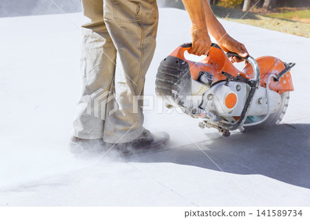 Masonry contractor is using concrete saw to cut through surface while dust clouds form around saw feet. 141589734