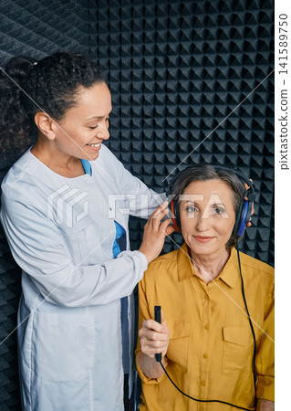 Audiometry. Audiologist doing hearing exam to a mature woman in soundproof audiometric cabin. Pure tone audiogram test Audiometry. Audiologist doing hearing exam to a mature woman in soundproof audiometric cabin. Pure tone audiogram test 141589750