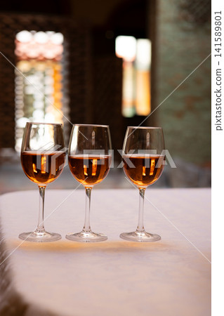 Three stemmed glasses filled with an amber-colored alcoholic beverage arranged on a restaurant table. Elegant still life with rose pink wine. Three stemmed glasses filled with an amber-colored alcoholic beverage arranged on a restaurant table. Elegant still life with rose pink wine. 141589801