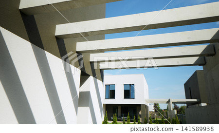 Modern concrete pergola creating strong shadow patterns on minimalist architectural facade under clear sky Modern concrete pergola creating strong shadow patterns on minimalist architectural facade under clear sky 141589938