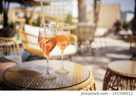 Two elegant glasses of orange cocktail with ice on a woven table in a sunny outdoor lounge area with comfortable seating and greenery in the background 141590012