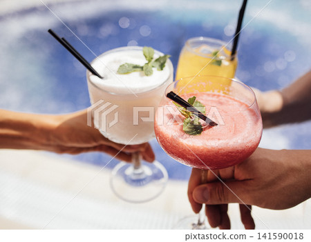 Three colorful cocktails with mint garnish held by hands, featuring a frozen strawberry drink, a yellow citrus drink, and a creamy white beverage near a pool Three colorful cocktails with mint garnish held by hands, featuring a frozen strawberry drink, a yellow citrus drink, and a creamy white beverage near a pool 141590018