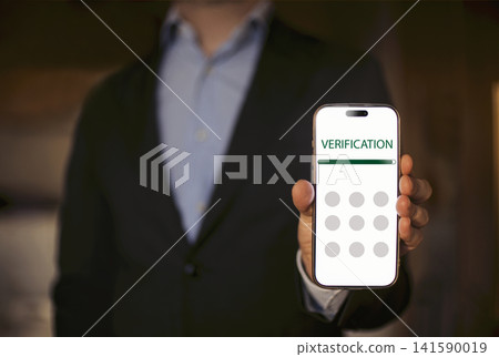 Man in black suit holding smartphone displaying verification screen with circular input fields, blurred background suggesting an indoor setting Man in black suit holding smartphone displaying verification screen with circular input fields, blurred background suggesting an indoor setting 141590019