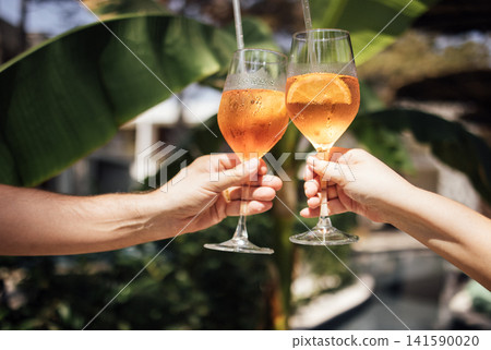Two hands clinking champagne glasses filled with orange beverage, surrounded by lush green tropical leaves in a bright outdoor setting Two hands clinking champagne glasses filled with orange beverage, surrounded by lush green tropical leaves in a bright outdoor setting 141590020