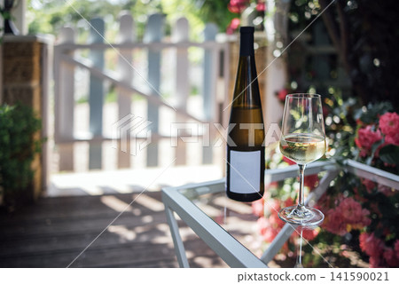 Bottle of white wine and glass on a table in a garden setting with a wooden gate and blooming flowers in the background Bottle of white wine and glass on a table in a garden setting with a wooden gate and blooming flowers in the background 141590021