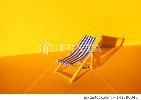 Miniature wooden deck chair with blue and white stripes positioned on a vibrant yellow background, casting a shadow, ideal for summer-themed designs and decorations Miniature wooden deck chair with blue and white stripes positioned on a vibrant yellow background, casting a shadow, ideal for summer-themed designs and decorations 141590045