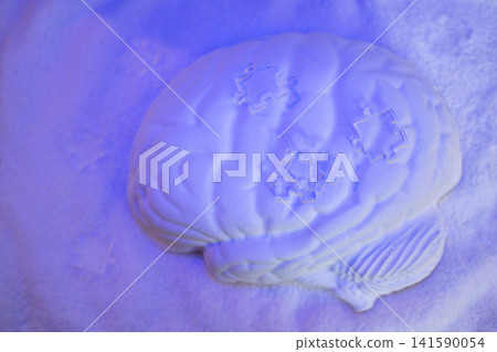 White brain sculpture placed on a textured surface with blue lighting, featuring puzzle piece designs embedded on the brain's surface White brain sculpture placed on a textured surface with blue lighting, featuring puzzle piece designs embedded on the brain's surface 141590054