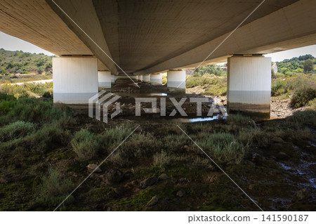 Underneath a concrete bridge with pillars over a dry riverbed and vegetation Arade river Portimao Underneath a concrete bridge with pillars over a dry riverbed and vegetation Arade river Portimao 141590187