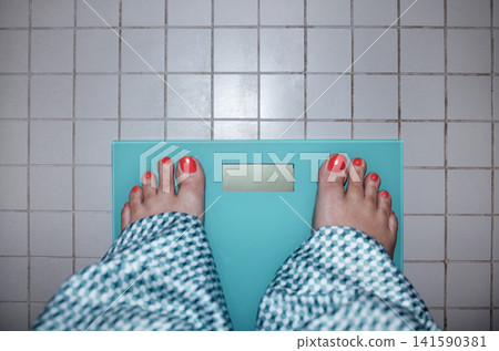 Bare feet with bright red nail polish standing on a turquoise bathroom scale on tiled floor, checkered bathrobe visible around ankles in a domestic setting 141590381