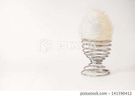 White egg-shaped object resting on a metal spiral egg holder against a plain light background, showcasing a minimalist design and modern aesthetic 141590412