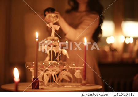 Female figure in soft focus arranging a tiered glass dessert stand with candles on a table, warm ambient lighting creating a cozy atmosphere in the background Female figure in soft focus arranging a tiered glass dessert stand with candles on a table, warm ambient lighting creating a cozy atmosphere in the background 141590413
