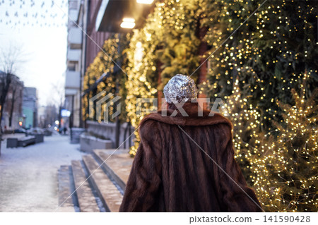 Woman in a brown fur coat walks along a snowy street adorned with festive lights and greenery, creating a winter holiday atmosphere in an urban setting Woman in a brown fur coat walks along a snowy street adorned with festive lights and greenery, creating a winter holiday atmosphere in an urban setting 141590428
