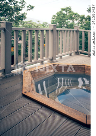 Octagonal wooden hot tub filled with clear water on a wooden deck surrounded by green trees and a railing in a tranquil outdoor setting Octagonal wooden hot tub filled with clear water on a wooden deck surrounded by green trees and a railing in a tranquil outdoor setting 141590437