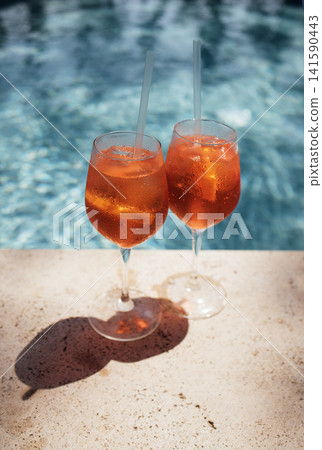 Two glasses of refreshing orange cocktails with ice and straws placed on a stone surface beside a sparkling blue swimming pool under bright sunlight Two glasses of refreshing orange cocktails with ice and straws placed on a stone surface beside a sparkling blue swimming pool under bright sunlight 141590443