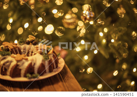 Holiday bundt cake decorated with icing and gingerbread cookies on a wooden table, with a blurred Christmas tree adorned with lights in the background Holiday bundt cake decorated with icing and gingerbread cookies on a wooden table, with a blurred Christmas tree adorned with lights in the background 141590451